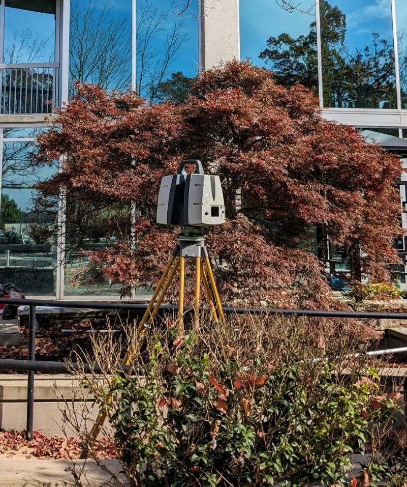 Closeup of a survey mapping camera on a tripod sitting in some bushes beside a city sidewalk.
