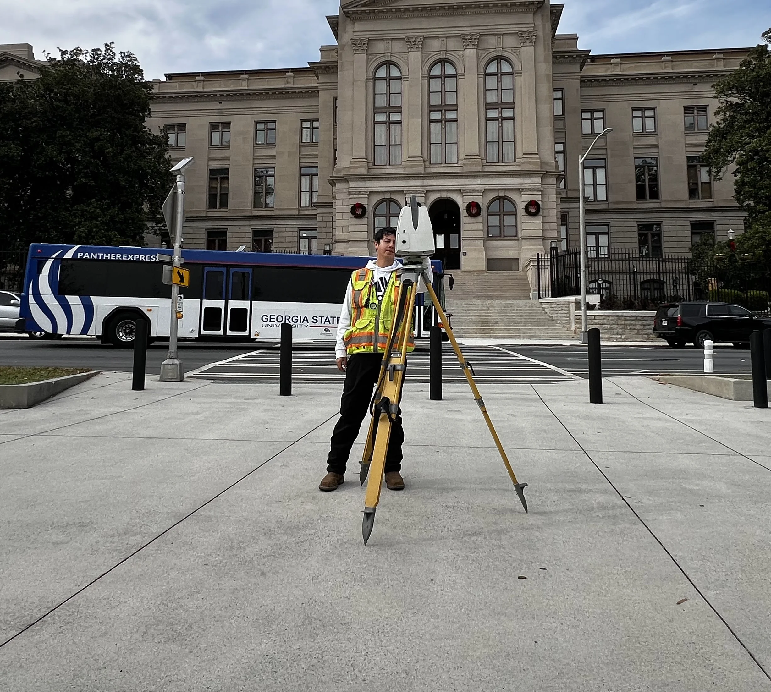 Surveyor standing behind a survey mapping camera on a city sidewalk. Behind them, a city bus passes and a large city building can be seen.