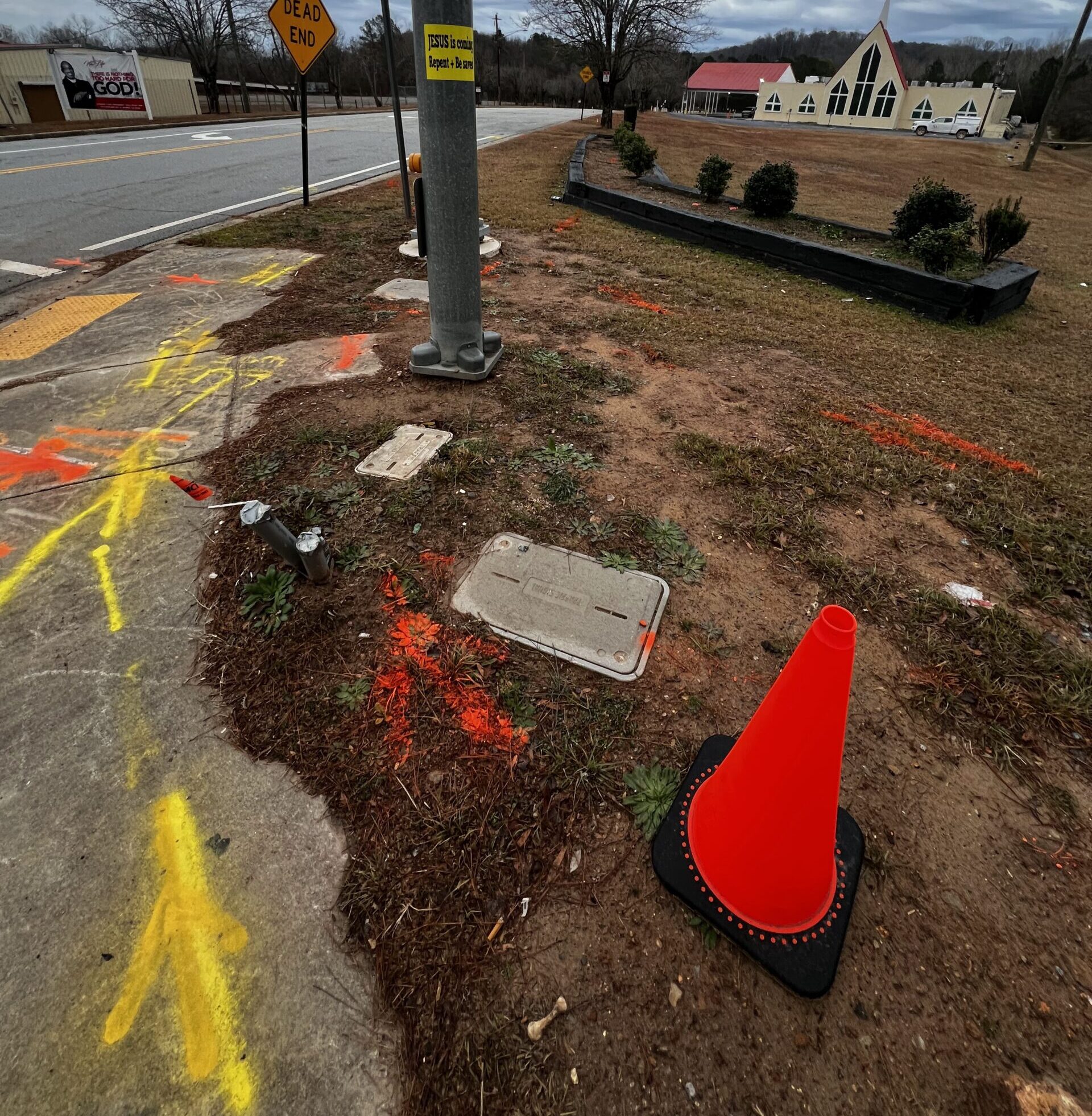 A residential street curb with an orange traffic cone, various yellow and orange spray-painted arrows and lines marking future construction.