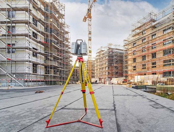 A survey mapping camera on a tripod sitting on a city street in front of a large commercial construction site.