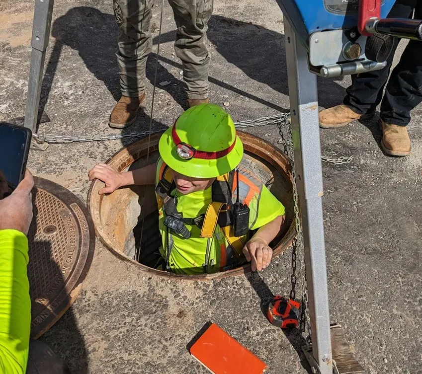 A surveyor dressed in reflective protective gear and hardhat lowering into a manhole.