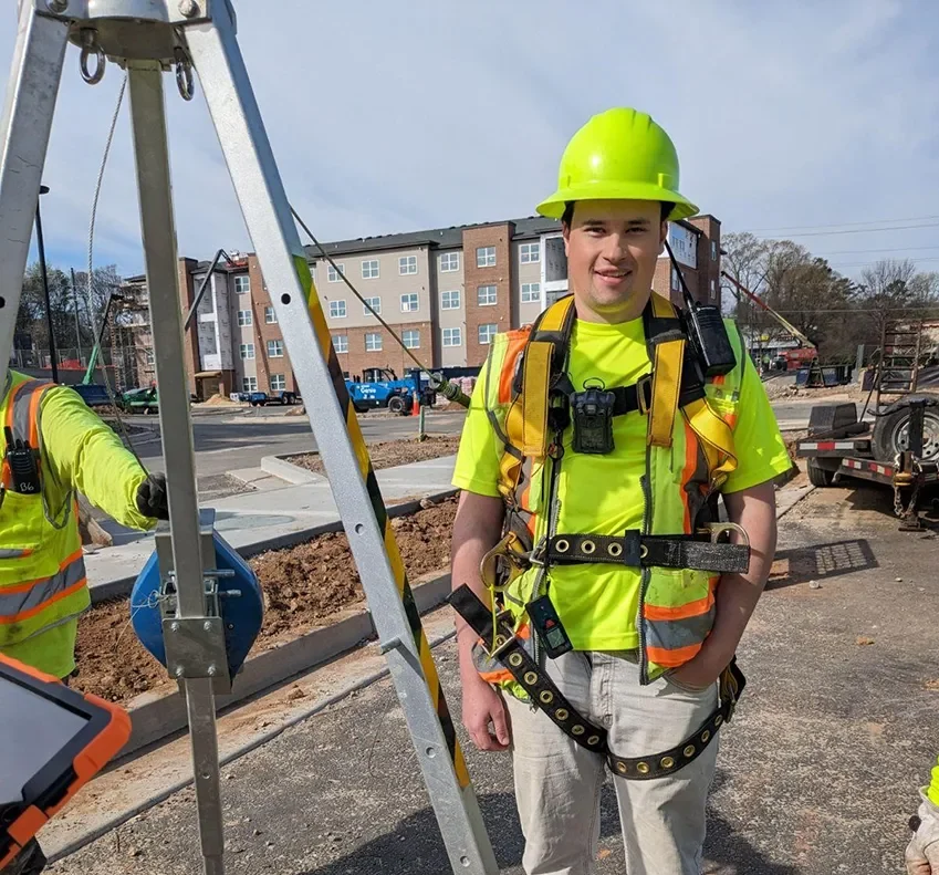 A surveyor dressed in reflective protective gear and hardhat smiling and standing beside a survey mapping camera tripod.